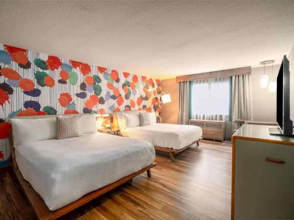 The image shows a hotel room with two beds, colorful wall art, wooden floor, a flat-screen TV, and a window with curtains.