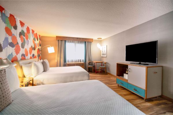 A hotel room with two beds, colorful wall, flat-screen TV, and a small table by the window.