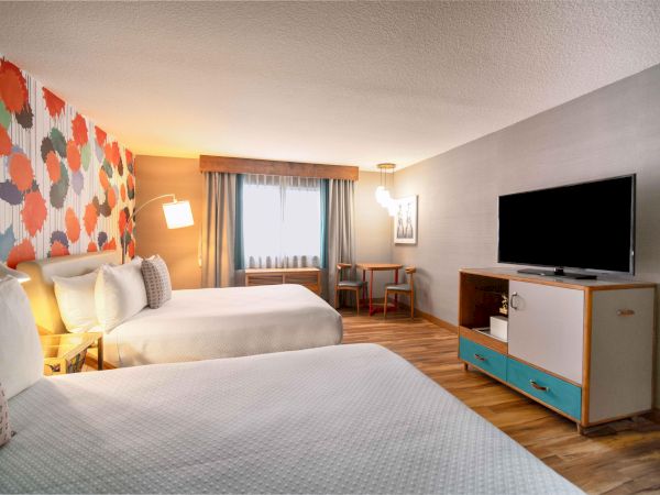 A hotel room with two beds, colorful wall, flat-screen TV, and a small table by the window.