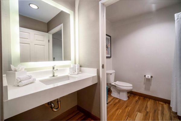 The image shows a modern bathroom with a lit vanity mirror, sink, and towels. There is a toilet and a shower curtain in the background.