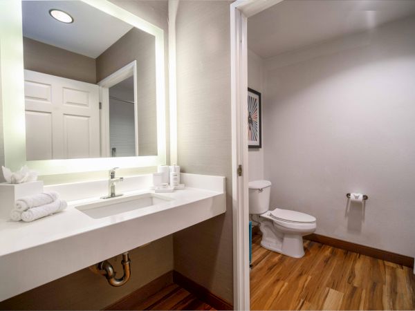 The image shows a modern bathroom with a lit vanity mirror, sink, and towels. There is a toilet and a shower curtain in the background.