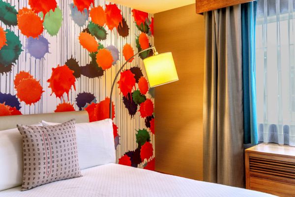 A colorful hotel room features a bed, patterned wall with abstract art, a floor lamp, pillow, and curtains by a window.