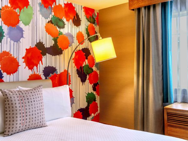 A colorful hotel room features a bed, patterned wall with abstract art, a floor lamp, pillow, and curtains by a window.