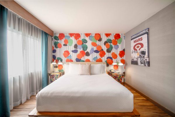 A brightly decorated bedroom with a colorful wall, large bed, two side tables, and a poster on the wall, well-lit by natural light.