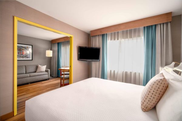 A hotel room with a bed, TV, and seating area; blue curtains and a framed art piece enhance the decor, creating a cozy atmosphere.