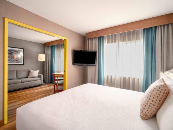 A hotel room with a bed, TV, and seating area; blue curtains and a framed art piece enhance the decor, creating a cozy atmosphere.