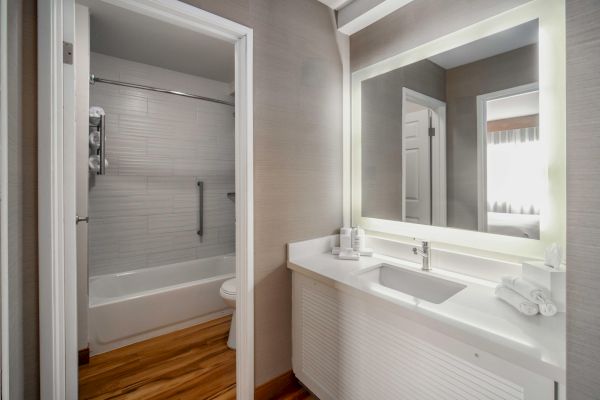 The image shows a modern bathroom with a bathtub, a shower, a large illuminated mirror, a sink, and neatly arranged towels.