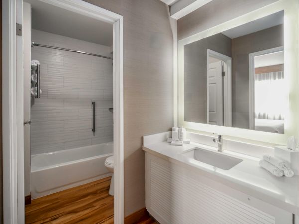 The image shows a modern bathroom with a bathtub, a shower, a large illuminated mirror, a sink, and neatly arranged towels.