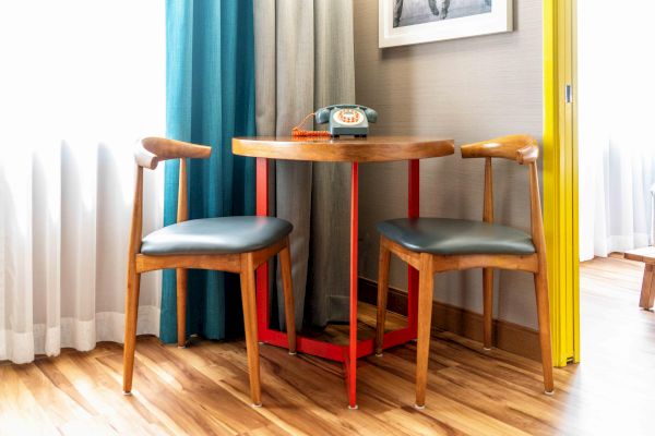 A small wooden table with two chairs, a retro telephone on top, near colorful curtains and a yellow wall, on wooden flooring.