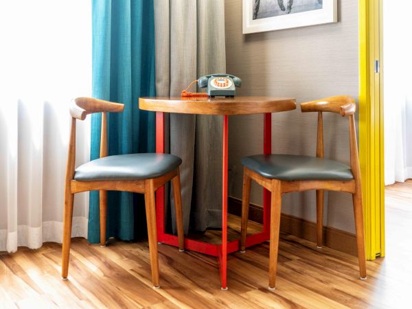 A small wooden table with two chairs, a retro telephone on top, near colorful curtains and a yellow wall, on wooden flooring.