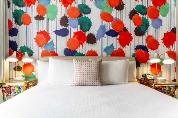 A bed with white linens is set against a vibrant, colorful splatter-paint wall. Two bedside tables hold modern lamps and decorations.