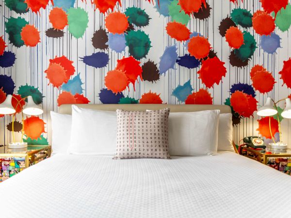A bed with white linens is set against a vibrant, colorful splatter-paint wall. Two bedside tables hold modern lamps and decorations.
