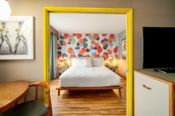 The image shows a modern hotel room with a bed, colorful wallpaper, wooden floor, and a TV. There's a table and chair in the foreground.