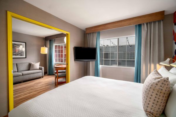 A modern hotel room with a bed, TV, window, and a separate seating area featuring a sofa and desk.
