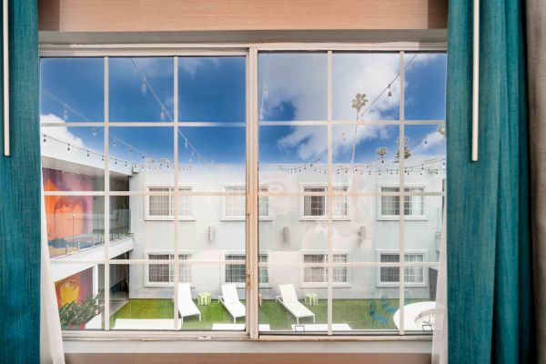 View through a window with teal curtains shows a courtyard, outdoor lounge chairs, and string lights against a blue sky with clouds.