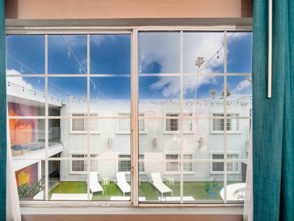 View through a window with teal curtains shows a courtyard, outdoor lounge chairs, and string lights against a blue sky with clouds.
