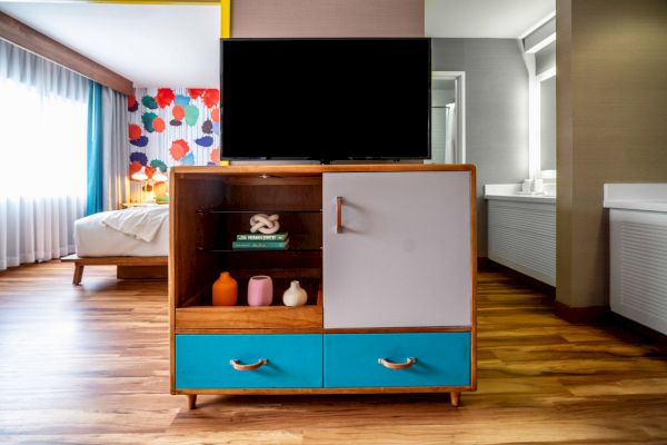 A hotel room features modern decor, a TV on a stand with colorful drawers, a partial view of the bed, and vibrant wallpaper.
