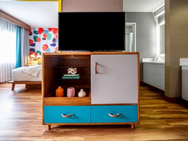 A hotel room features modern decor, a TV on a stand with colorful drawers, a partial view of the bed, and vibrant wallpaper.