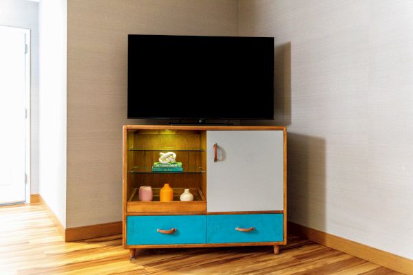 A TV on a wooden stand with blue drawers, a closed cabinet, and shelves holding folded towels and small bottles in a corner room.
