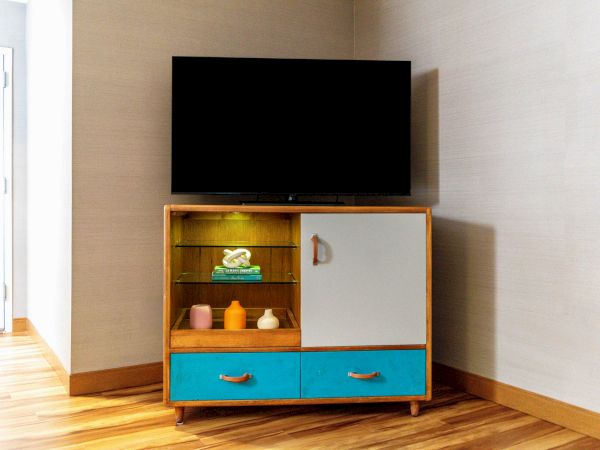 A TV on a wooden stand with blue drawers, a closed cabinet, and shelves holding folded towels and small bottles in a corner room.