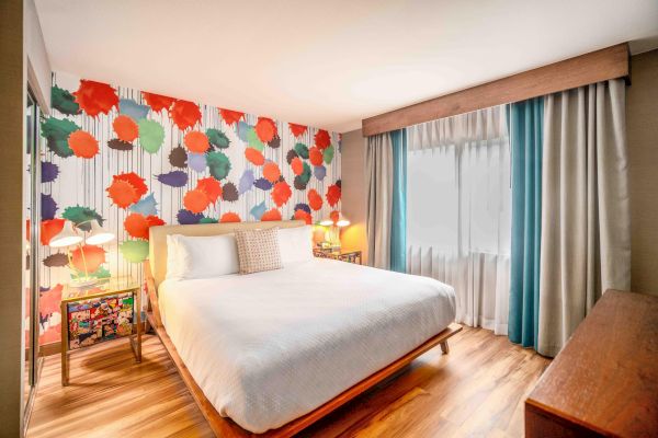 A brightly decorated bedroom with a colorful floral wall, a white bed, wooden floor, window with curtains, and bedside tables with lamps.