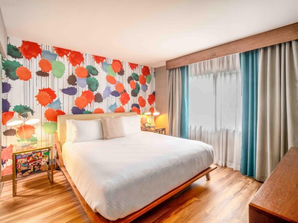A brightly decorated bedroom with a colorful floral wall, a white bed, wooden floor, window with curtains, and bedside tables with lamps.