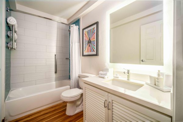 A modern bathroom with a white bathtub, shower curtain, toilet, sink with mirror, and a colorful artwork on the wall.
