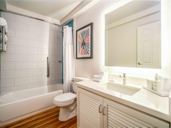 A modern bathroom with a white bathtub, shower curtain, toilet, sink with mirror, and a colorful artwork on the wall.
