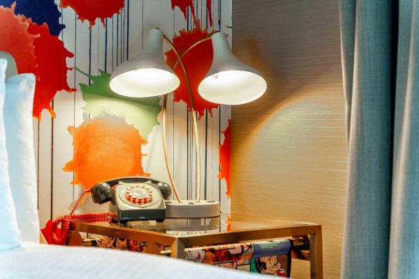 A retro phone is on a table beside two lamps, with a colorful paint splatter wallpaper behind it.