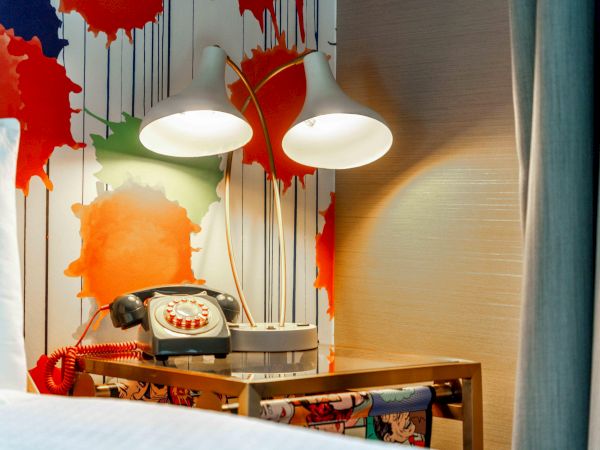 A retro phone is on a table beside two lamps, with a colorful paint splatter wallpaper behind it.