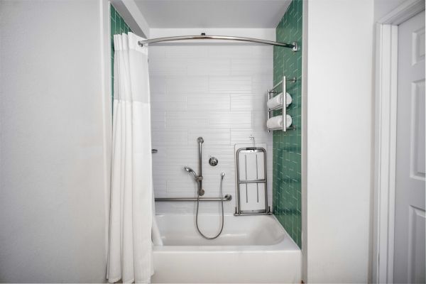 This image shows a bathroom with a bathtub, white curtain, green tile walls, showerhead, safety rails, and towel rack, ensuring accessibility.