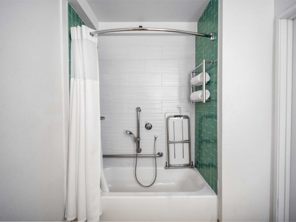 This image shows a bathroom with a bathtub, white curtain, green tile walls, showerhead, safety rails, and towel rack, ensuring accessibility.