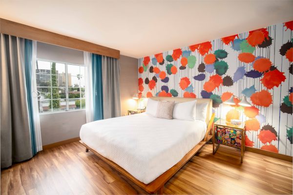 A brightly decorated bedroom with a colorful accent wall, a large bed, hardwood floor, lamps, and a window with curtains.