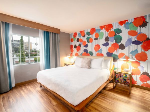 A brightly decorated bedroom with a colorful accent wall, a large bed, hardwood floor, lamps, and a window with curtains.