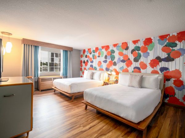 The image shows a modern hotel room with two beds, wood flooring, and a vibrant, colorful wall design featuring large abstract patterns.