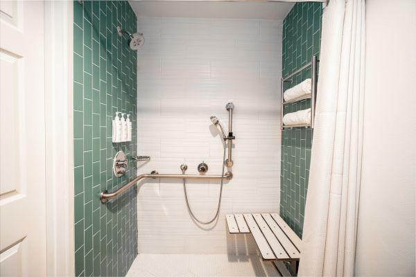 This image shows an accessible shower with green and white tiles, a handrail, a showerhead, a foldable bench, and toiletries on the wall.