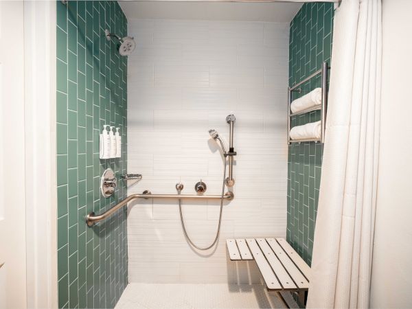 This image shows an accessible shower with green and white tiles, a handrail, a showerhead, a foldable bench, and toiletries on the wall.