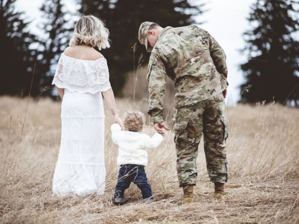 A family, with a woman in a white dress, a person in military uniform, and a child, holds hands while walking in a grassy area.