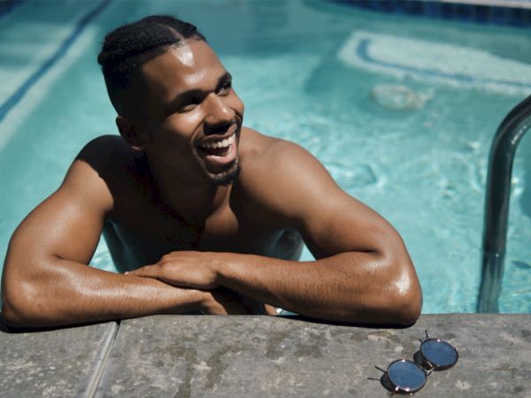 A person is smiling while leaning on the edge of a swimming pool, with sunglasses resting nearby on the poolside.
