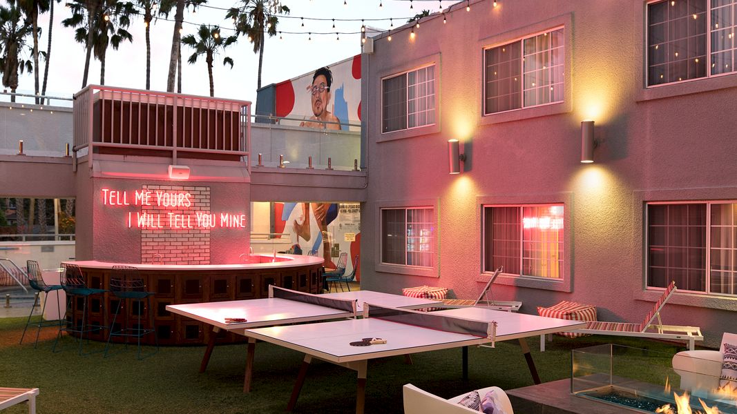 Cozy outdoor lounge with ping pong tables, string lights, and a neon sign. Comfy seating and a fire pit create a relaxing atmosphere.