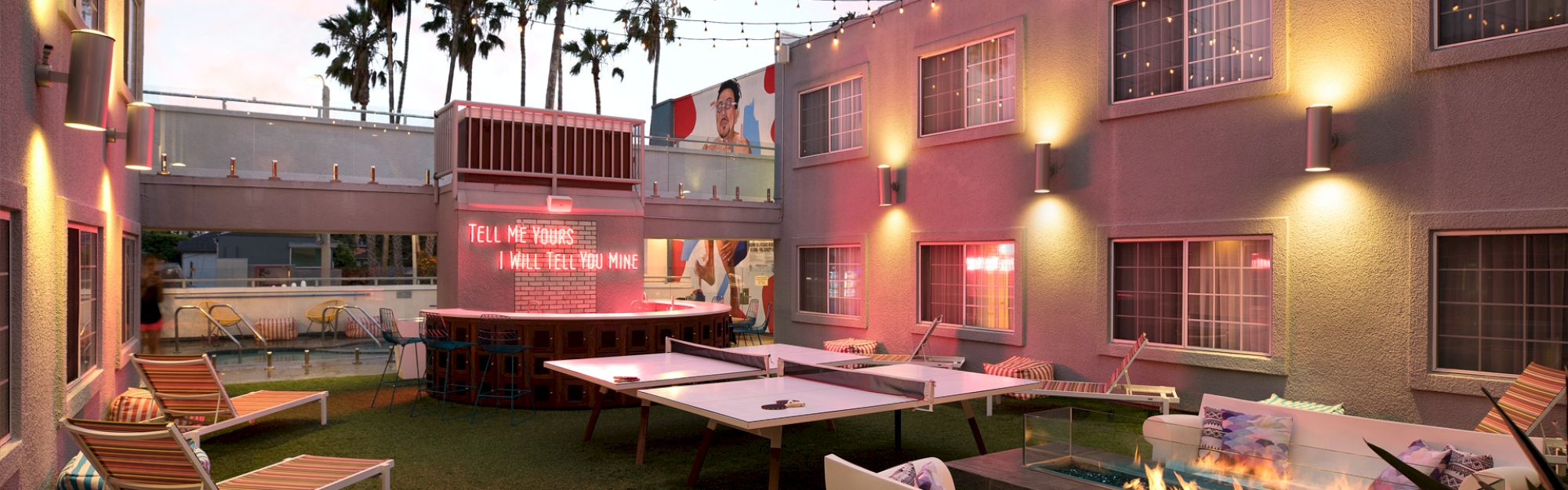Cozy outdoor lounge with ping pong tables, string lights, and a neon sign. Comfy seating and a fire pit create a relaxing atmosphere.