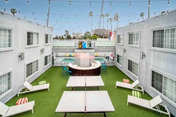 An outdoor courtyard with lounge chairs, a ping pong table, string lights, and a bar in a modern setting, surrounded by buildings.