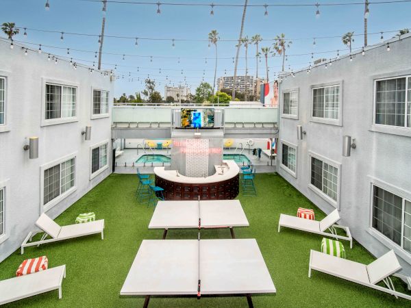 An outdoor courtyard with lounge chairs, a ping pong table, string lights, and a bar in a modern setting, surrounded by buildings.