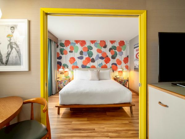 A modern bedroom with colorful decor, featuring a bed, wood floor, a pop art-style wall, and framed art on a beige wall.
