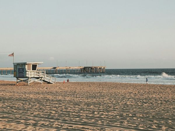 A beach scene with a lifeguard tower, people on the sand, a pier extending into the ocean, and a clear blue sky overhead.