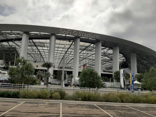 The image shows a large stadium with the name "SoFi Stadium" on its structure, surrounded by greenery and a partly cloudy sky.