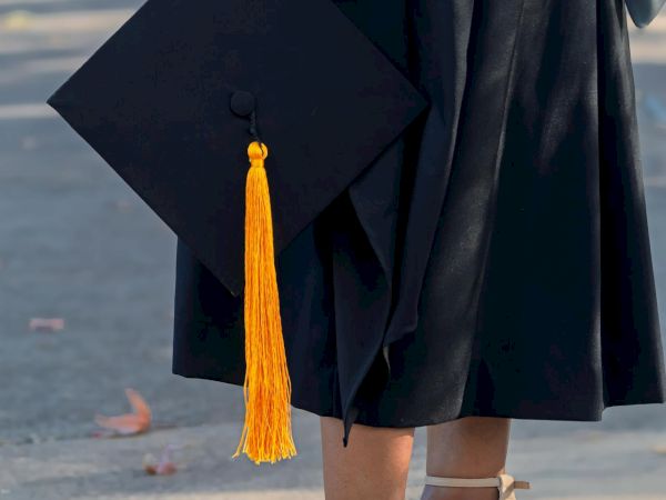A person in a graduation gown holds a cap with a yellow tassel, wearing beige high heels.