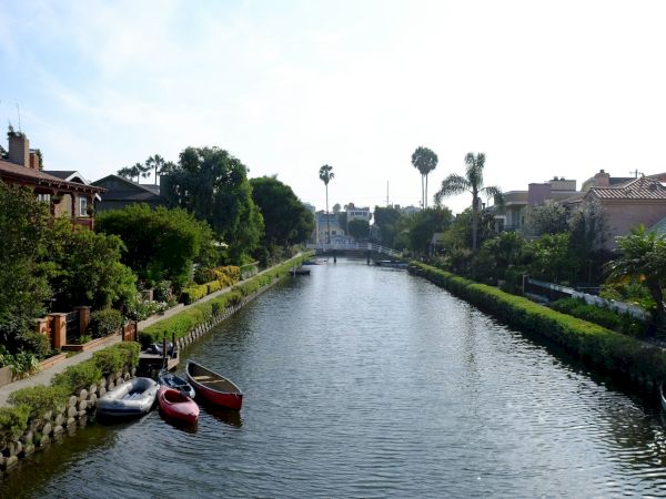 A serene canal lined with lush greenery and houses, small boats moored on the side, and palm trees under a clear, blue sky.