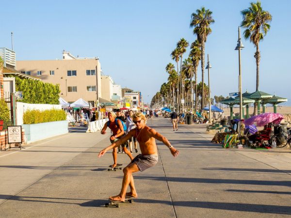 People are skateboarding on a beachside boardwalk with palm trees and various stalls in the background.