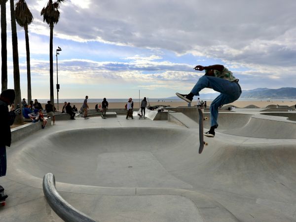 A skateboarder performs a trick in a skatepark by the beach, with people and palm trees in the background under a cloudy sky.